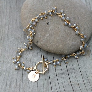 Könnte beinhalten: Ein zartes, goldfarbenes Armband mit kleinen, grauen Edelsteinperlen. Das Armband hat einen Knebelverschluss und einen kleinen, runden Anhänger mit dem eingravierten Buchstaben "C". Das Armband wird auf einem grauen Stein präsentiert.