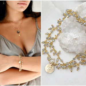 May include: A gold-coloured jewellery set featuring a necklace with a square pendant and a charm, and a delicate bracelet with small charms. A second bracelet with grey beads and a gold clasp is also shown. The jewellery is displayed on a white background.
