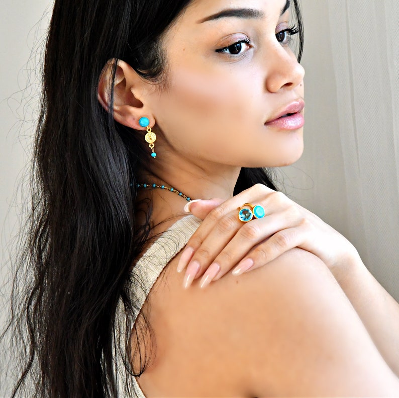 May include: A collection of turquoise and gold jewelry. The set includes a pair of turquoise stud earrings, a turquoise and gold pendant necklace, and a gold ring with two turquoise stones. The jewelry is displayed on a person.