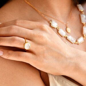 May include: A gold ring with a round, white gemstone is displayed on a hand. A gold necklace with rectangular and diamond-shaped white gemstones is also visible. The jewelry is set against a neutral background.