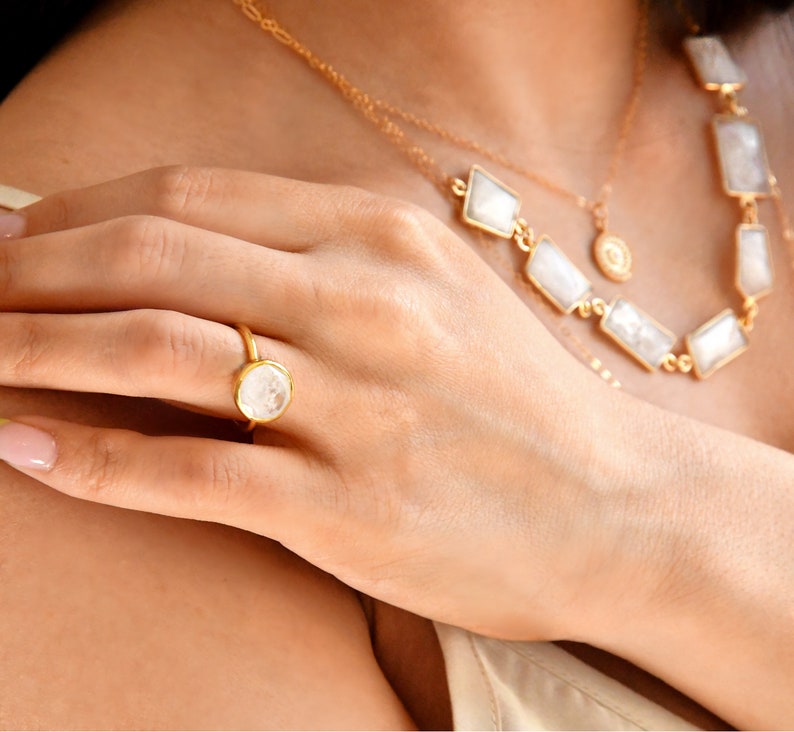 May include: A gold ring with a round, white gemstone is displayed on a hand. A gold necklace with rectangular and diamond-shaped white gemstones is also visible. The jewelry is set against a neutral background.
