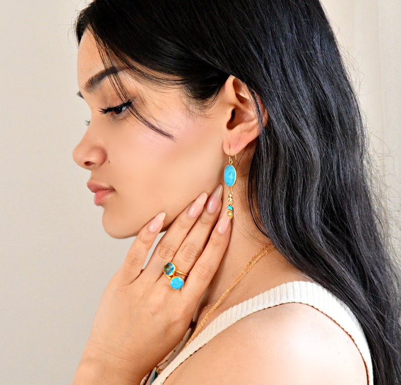 May include: Gold-toned jewelry set featuring a ring and earrings with turquoise-colored stones. The ring has two stones, and the earrings are long with a single oval stone and dangling accents. The jewelry is worn by a person.