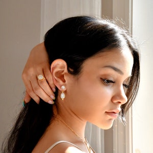 May include: A gold ring and earrings set featuring a pearlescent stone. The ring has a round stone set in a gold band. The earrings have a pearl stud with a teardrop-shaped pearl pendant. The jewelry is worn by a person.