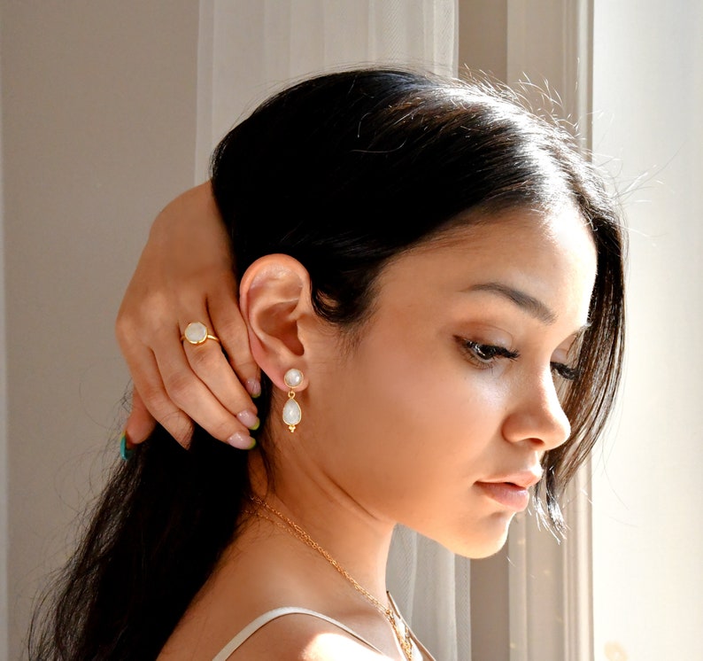 May include: A gold ring and earrings set featuring a pearlescent stone. The ring has a round stone set in a gold band. The earrings have a pearl stud with a teardrop-shaped pearl pendant. The jewelry is worn by a person.