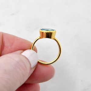 May include: A gold ring with a large, oval-shaped, light blue gemstone. The ring's band and setting are a bright gold color. The gemstone appears to be smooth and polished, reflecting light. The ring is held by a person.