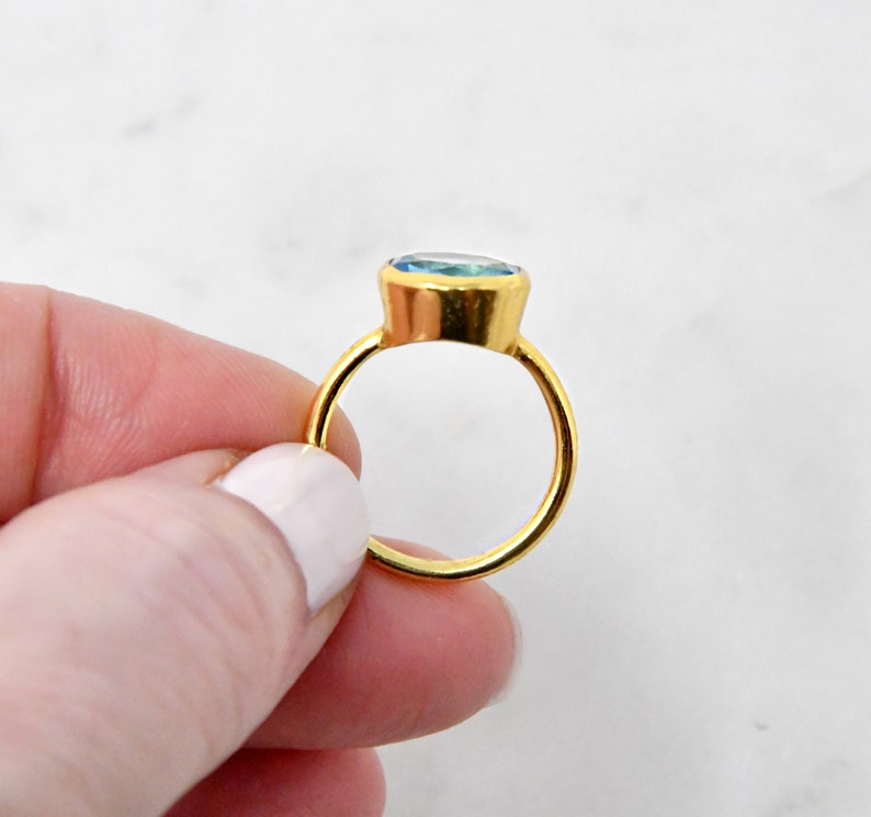 May include: A gold ring with a large, oval-shaped, light blue gemstone. The ring's band and setting are a bright gold color. The gemstone appears to be smooth and polished, reflecting light. The ring is held by a person.