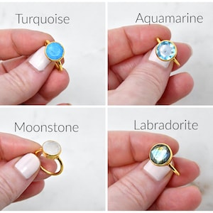 May include: Four gold-colored rings with different gemstones: turquoise, aquamarine, moonstone, and labradorite. Each ring features a round gemstone set in a simple gold band. The text "Turquoise", "Aquamarine", "Moonstone", and "Labradorite" is displayed above each ring.