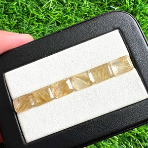 Pode incluir: Uma fileira de seis pedras preciosas de quartzo rutilado dourado, cada uma cortada em forma retangular. As pedras são exibidas em uma superfície branca dentro de uma caixa retangular preta, contra um fundo de grama verde.