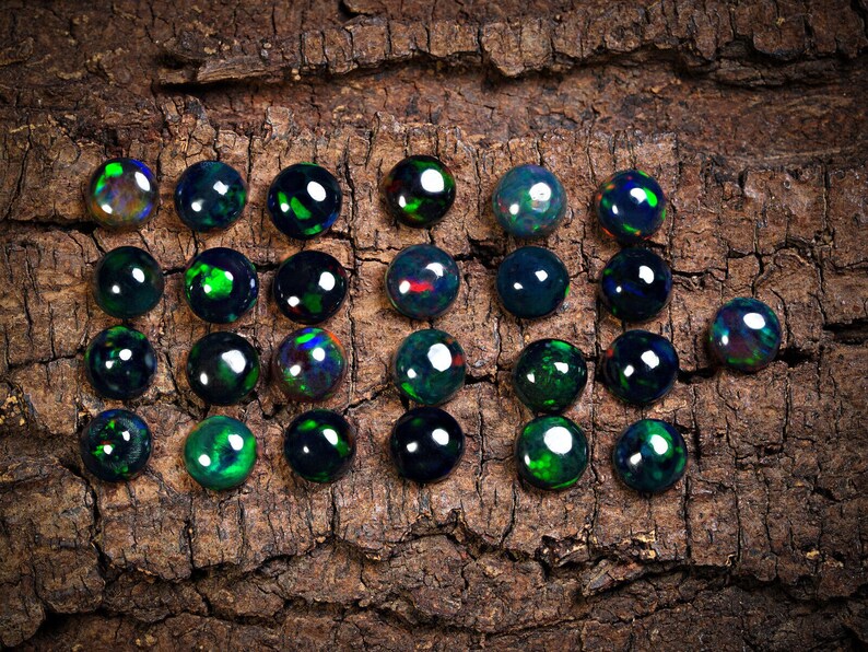 Puede incluir: Una colecci&oacute;n de 20 cabujones de &oacute;palo negro, cada uno con un brillo iridiscente verde y azul &uacute;nico, dispuestos sobre un trozo de corteza.