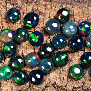 Puede incluir: Una colecci&oacute;n de piedras preciosas de &oacute;palo negro con destellos iridiscentes verdes, azules y rojos. Los &oacute;palos son redondos y lisos, y est&aacute;n dispuestos en un patr&oacute;n aleatorio sobre una pieza de madera.