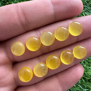 May include: A collection of ten round, polished, yellow gemstones. The stones are smooth and have a translucent quality, with a warm, golden hue. The gemstones are displayed against a background of green grass.