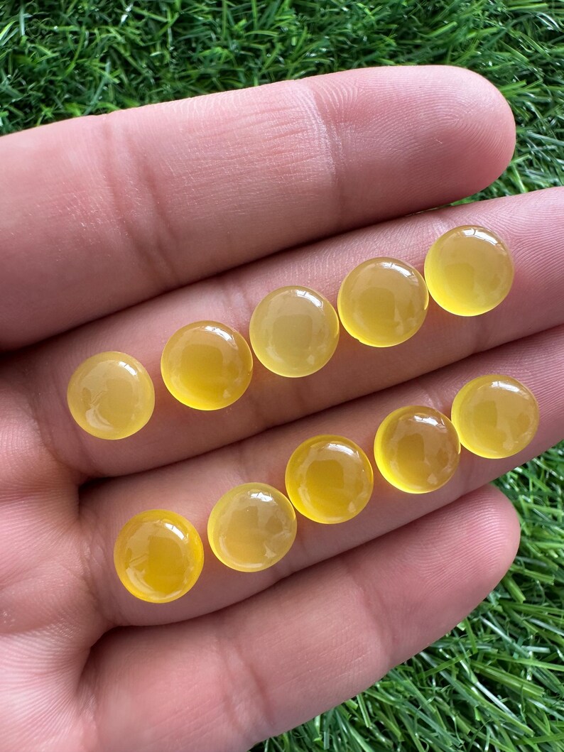 May include: A collection of ten round, polished, yellow gemstones. The stones are smooth and have a translucent quality, with a warm, golden hue. The gemstones are displayed against a background of green grass.