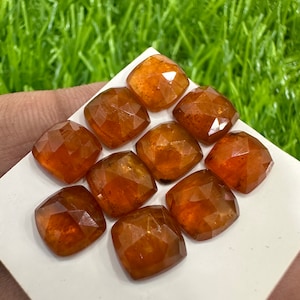 May include: Ten faceted, orange-brown gemstones, cut in a square shape, are arranged on a white surface. The gemstones are all approximately the same size and have a slightly translucent appearance.