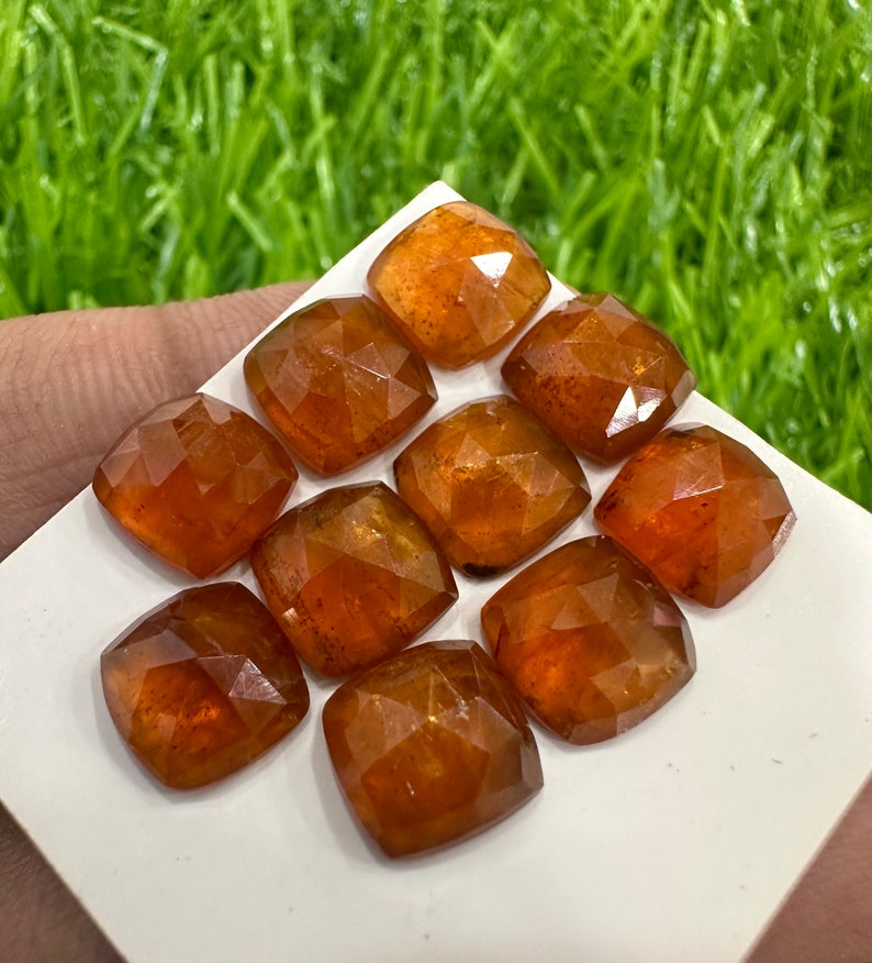 May include: Ten faceted, orange-brown gemstones, cut in a square shape, are arranged on a white surface. The gemstones are all approximately the same size and have a slightly translucent appearance.