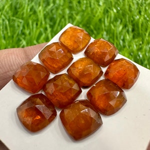 May include: Ten faceted, orange-brown gemstones, each with a square shape, are arranged on a white surface. The gemstones are cut with many facets, giving them a sparkling appearance.