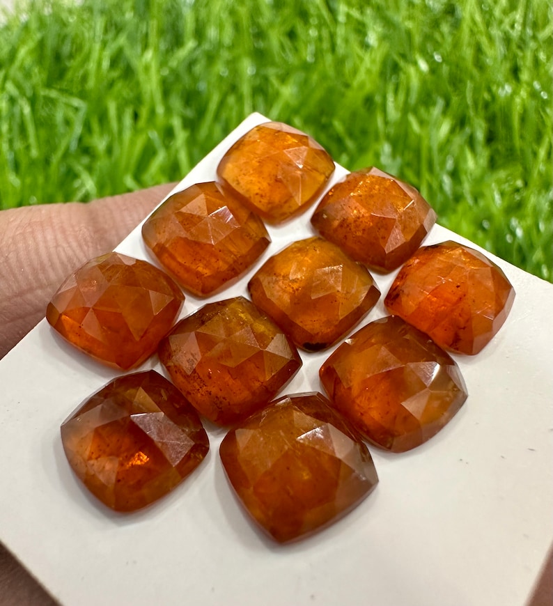 May include: Ten faceted, orange-brown gemstones, each with a square shape, are arranged on a white surface. The gemstones are cut with many facets, giving them a sparkling appearance.