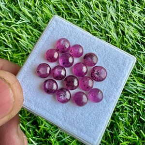 May include: A collection of 14 round, pink gemstones with black flecks. The gemstones are displayed on a white surface.
