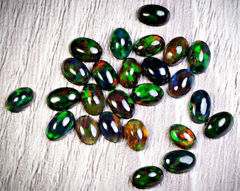 May include: A collection of oval-shaped black opal gemstones with vibrant flashes of green, red, and orange. The opals are polished and display a play-of-color effect, reflecting light. The gemstones are scattered on a white, textured surface.