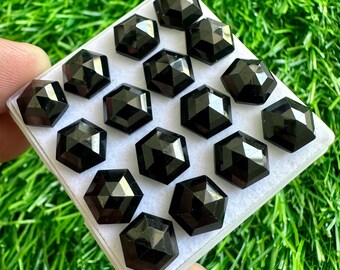 May include: A close-up of a white square box containing 15 black gemstone cabochons. The cabochons are faceted and have a hexagonal shape. The box is sitting on a green grassy surface.