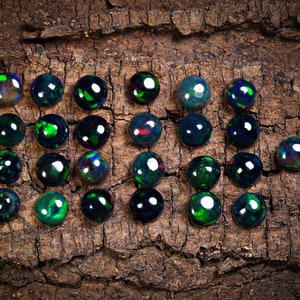 Puede incluir: Una colecci&oacute;n de 20 cabujones de &oacute;palo negro, cada uno con un brillo iridiscente verde y azul &uacute;nico, dispuestos sobre un trozo de corteza.