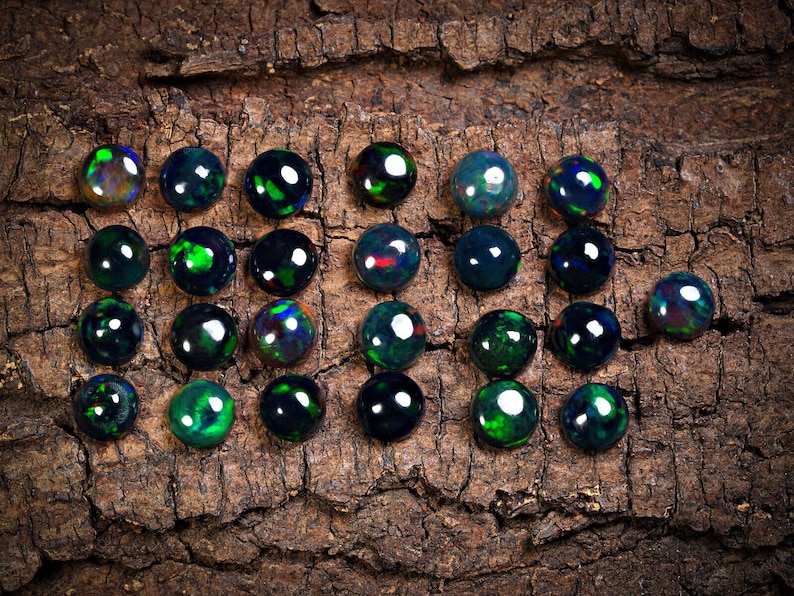 Puede incluir: Una colecci&oacute;n de 20 cabujones de &oacute;palo negro, cada uno con un brillo iridiscente verde y azul &uacute;nico, dispuestos sobre un trozo de corteza.