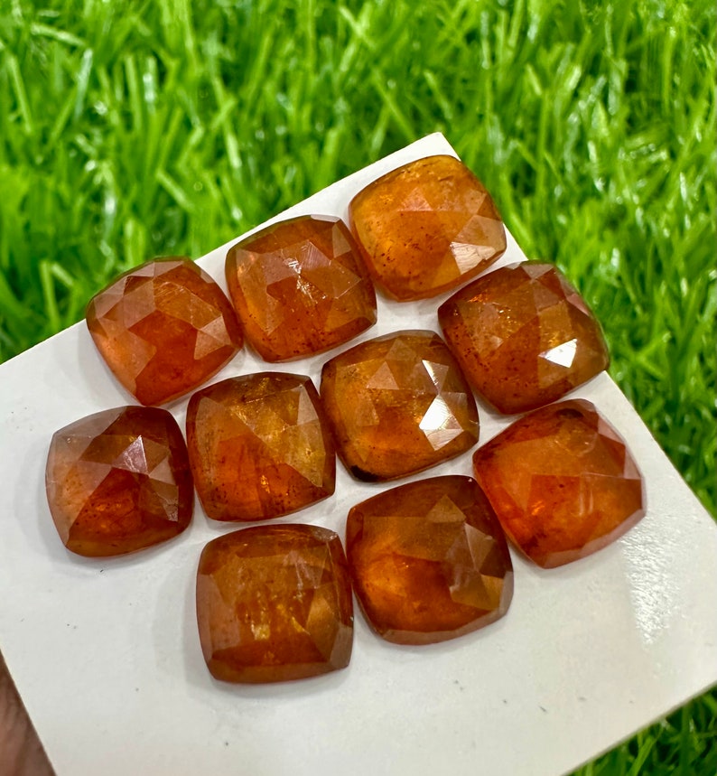 May include: Ten loose, faceted, orange-brown gemstones. The gemstones are cut in a cushion shape and have a glossy finish.