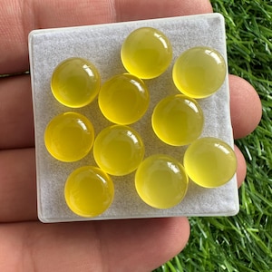 May include: A collection of ten round, yellow gemstones displayed on a square, light gray surface. The gemstones have a smooth, polished appearance and a translucent quality. The background is a vibrant green, suggesting a natural setting.