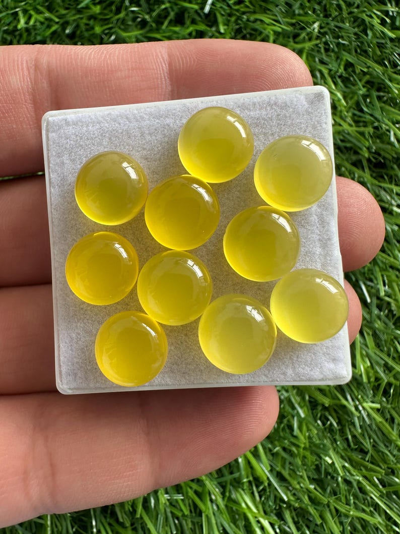 May include: A collection of ten round, yellow gemstones displayed on a square, light gray surface. The gemstones have a smooth, polished appearance and a translucent quality. The background is a vibrant green, suggesting a natural setting.