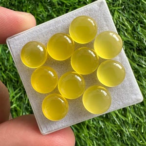 May include: A collection of ten round, translucent yellow gemstones displayed on a light gray surface. The gemstones have a polished, glossy appearance, and are set against a backdrop of green grass.