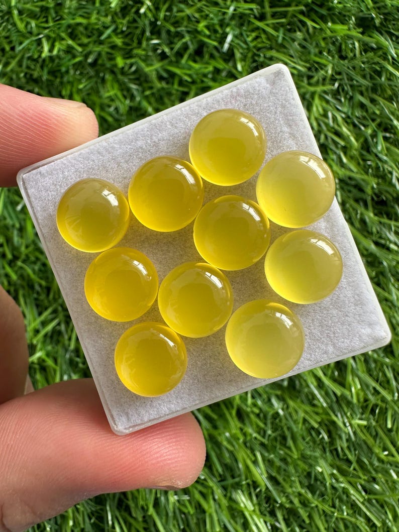 May include: A collection of ten round, translucent yellow gemstones displayed on a light gray surface. The gemstones have a polished, glossy appearance, and are set against a backdrop of green grass.