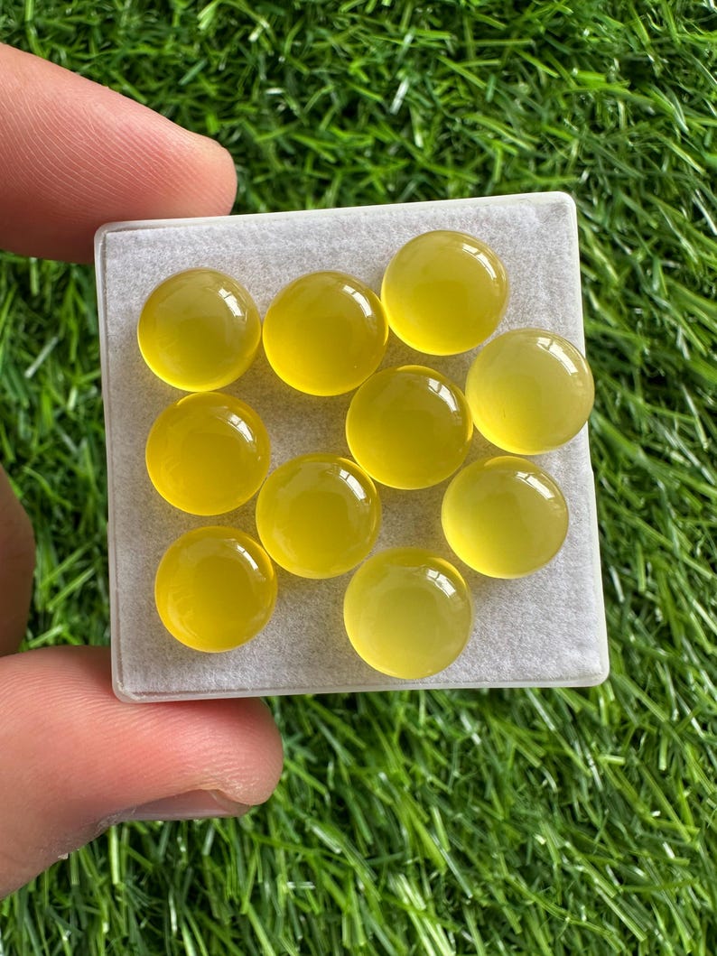 May include: A collection of ten round, yellow gemstones displayed in a clear plastic container. The gemstones have a smooth, polished surface and a translucent appearance. The container is held against a backdrop of green grass.
