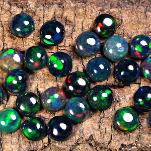 Puede incluir: Una colecci&oacute;n de piedras preciosas de &oacute;palo negro con destellos iridiscentes verdes, azules y rojos. Los &oacute;palos son redondos y lisos, y est&aacute;n dispuestos en un patr&oacute;n aleatorio sobre una pieza de madera.