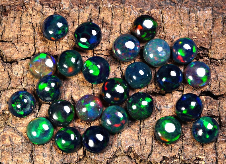 Puede incluir: Una colecci&oacute;n de piedras preciosas de &oacute;palo negro con destellos iridiscentes verdes, azules y rojos. Los &oacute;palos son redondos y lisos, y est&aacute;n dispuestos en un patr&oacute;n aleatorio sobre una pieza de madera.