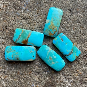 Puede incluir: Cinco piedras turquesa, cada una con un patrón único de azul, verde y marrón, están dispuestas sobre una superficie gris rugosa. Las piedras son de forma rectangular y tienen un acabado liso y pulido.