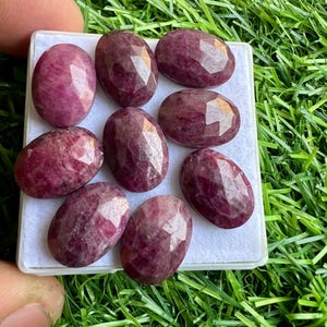 Puede incluir: Ocho piedras preciosas de rubí talladas en forma ovalada en varios tonos de rosa y rojo. Las piedras preciosas se muestran sobre una superficie blanca.
