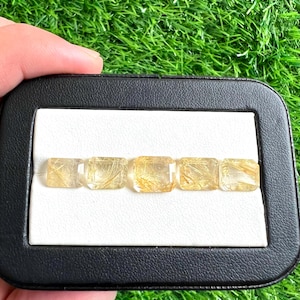 Pode incluir: Cinco pedras preciosas retangulares amarelo-douradas com inclusões douradas internas são exibidas em uma caixa preta com interior branco. As pedras são seguradas por uma mão, com um fundo verde gramado.