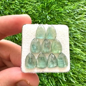 May include: Ten light blue-green gemstones, each cut into a faceted, teardrop shape. The gems are displayed on a white surface, set against green grass. The gems are held by a person's hand.