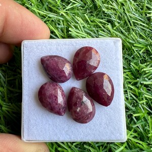 Puede incluir: Cinco piedras preciosas de rubí facetadas en forma de pera en una caja de terciopelo blanco. Los rubíes son de color rojo intenso y tienen un ligero tono púrpura.