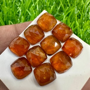 May include: Ten faceted, orange-brown gemstones, cut in a square shape, are displayed on a white surface. The gemstones are arranged in a staggered pattern.