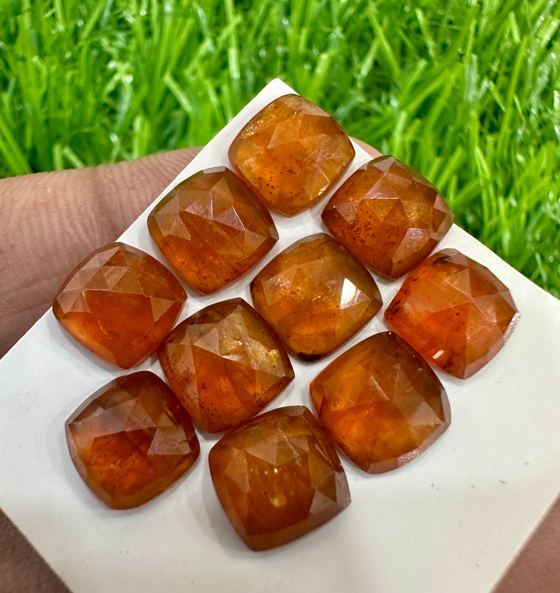 May include: Ten faceted, orange-brown gemstones, cut in a square shape, are displayed on a white surface. The gemstones are arranged in a staggered pattern.