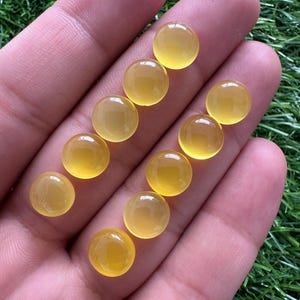 May include: A collection of ten round, polished, yellow gemstones. The gems are smooth and translucent, with a warm, golden hue. They are displayed on a hand against a backdrop of green grass. The gemstones are likely cabochons, suitable for jewelry making.