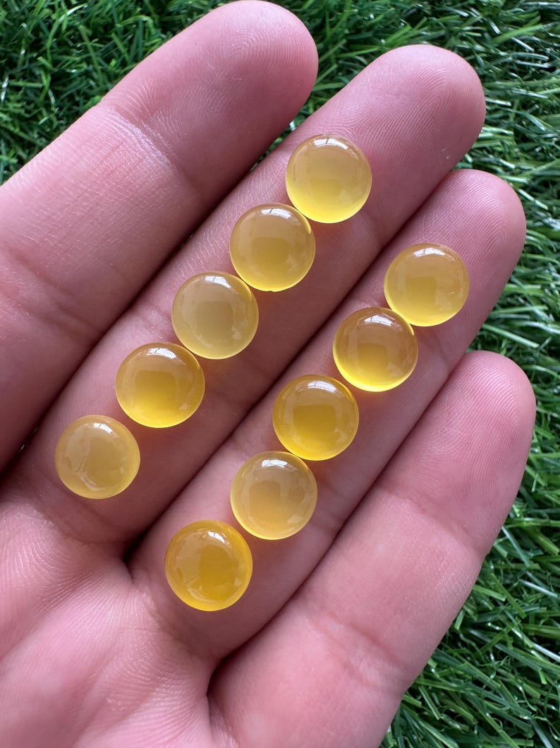 May include: A collection of ten round, polished, yellow gemstones. The gems are smooth and translucent, with a warm, golden hue. They are displayed on a hand against a backdrop of green grass. The gemstones are likely cabochons, suitable for jewelry making.