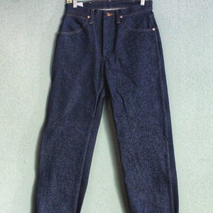 Puede incluir: Un par de pantalones vaqueros de mezclilla azul oscuro con dobladillo. Los pantalones vaqueros est&aacute;n colgados en una percha de madera.