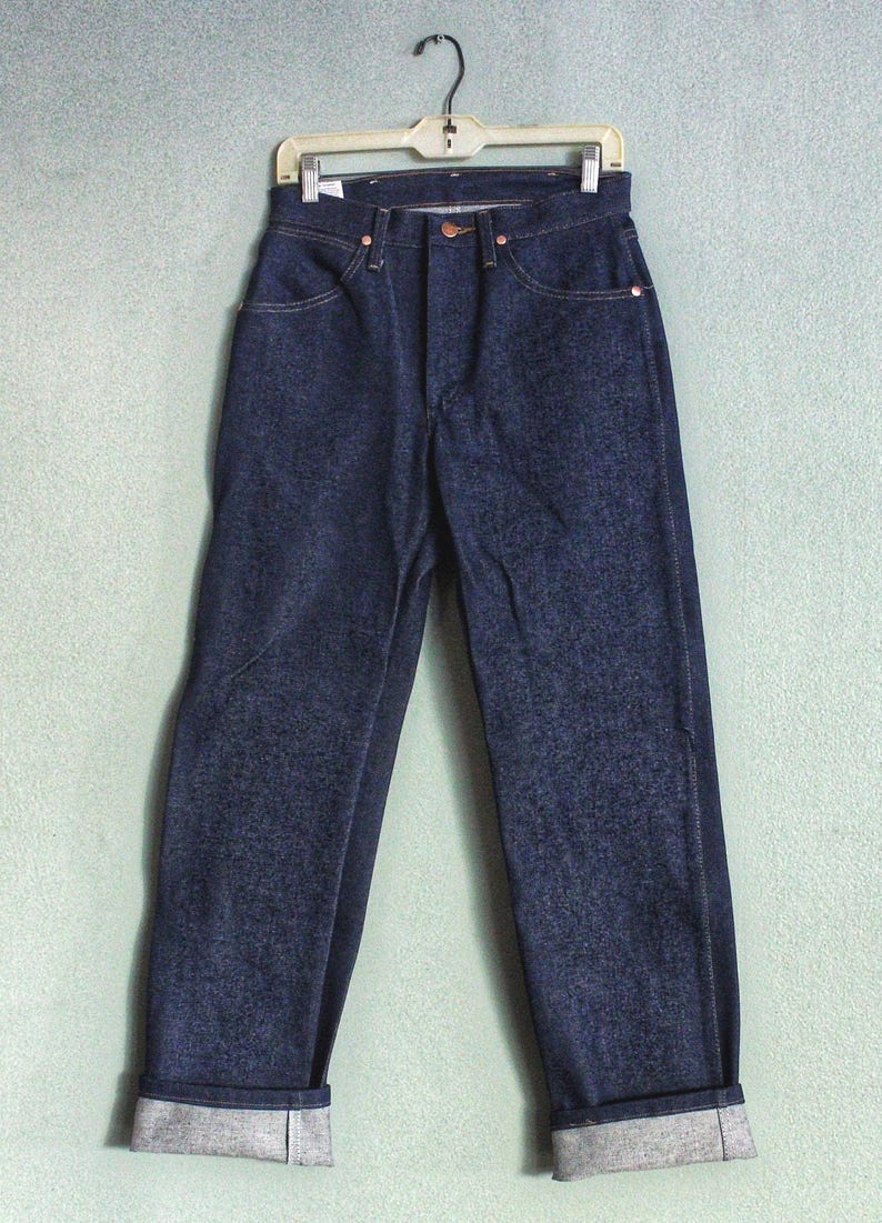 Puede incluir: Un par de pantalones vaqueros de mezclilla azul oscuro con dobladillo. Los pantalones vaqueros est&aacute;n colgados en una percha de madera.
