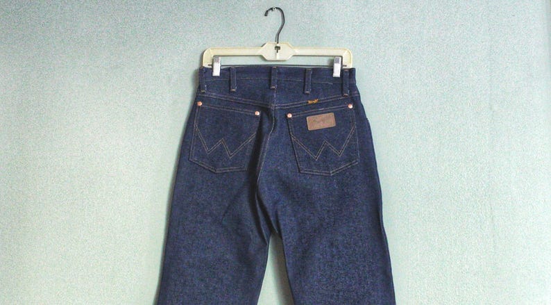 Puede incluir: Un par de pantalones vaqueros de mezclilla azul oscuro con un parche de cuero marr&oacute;n en el bolsillo trasero que dice "Wrangler".