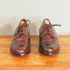May include: A pair of brown leather lace-up shoes with a polished finish. The shoes are on a wooden surface.
