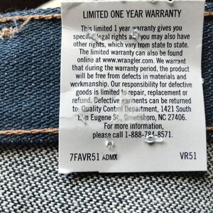 Puede incluir: Una etiqueta blanca con texto negro que detalla una garant&iacute;a limitada de un a&ntilde;o para los jeans Wrangler. El texto incluye los detalles de la garant&iacute;a, la informaci&oacute;n de contacto y la direcci&oacute;n del Departamento de Control de Calidad. La etiqueta tambi&eacute;n incluye el c&oacute;digo de producto 7FAVR51 ADMX y VR51.