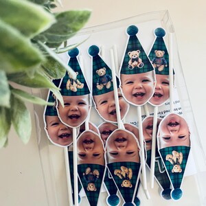 Puede incluir: Un conjunto de adornos para pasteles con la cara de un beb&eacute; sonriente sobre un gorro de fiesta a cuadros verdes y azules con un gr&aacute;fico de oso de peluche y un pomp&oacute;n azul oscuro, montados sobre palitos blancos.