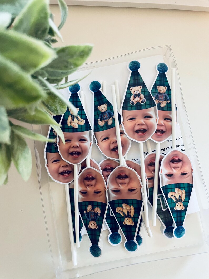 Puede incluir: Un conjunto de adornos para pasteles con la cara de un beb&eacute; sonriente sobre un gorro de fiesta a cuadros verdes y azules con un gr&aacute;fico de oso de peluche y un pomp&oacute;n azul oscuro, montados sobre palitos blancos.