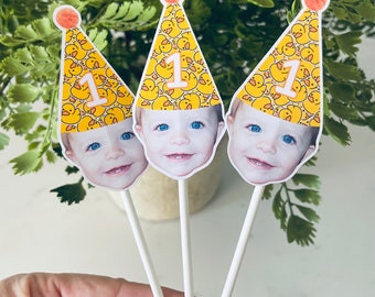 May include: Three yellow and orange party hats with a baby's face on each. The hats have a pattern of yellow rubber duckies and the number "1" on the front. The hats are on white sticks.